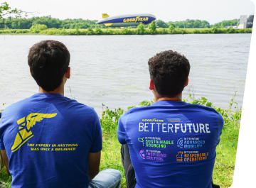 View from behind two Goodyear intern sitting next to each other with Goodyear t-shirts on in front of a lake with the Goodyear Blimp flying over the water in the distance